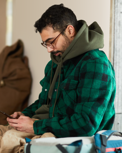 Man looking at smartphone in shelter. A man wearing a green jacket and hoodie sits on the floor of a shelter with a blanket and holding a smart phone. Others can be seen in the background sitting on the floor as well with bags, blankets, and eating food.