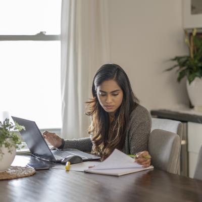 Young woman looking at papers and working on laptop.